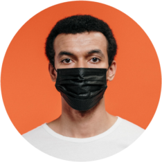 Male person of color wearing black medical face mask on orange background.